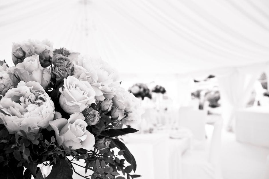 A floral arrangement featuring a large bouquet of fresh roses, peonies, and assorted blooms with delicate petals and lush green foliage, placed on a table under a white event marquee. The bouquet is w