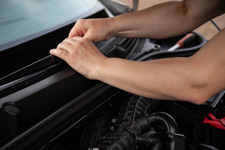Close-up of a person's hands opening the hood of a car to inspect the engine compartment, which contains various mechanical parts and hoses. The person has neatly trimmed nails and is outdoors with na