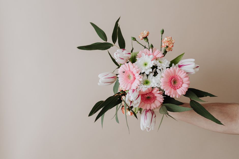 A hand holding a fresh floral bouquet featuring pink gerbera daisies, white daisies, and pink tulips, accented with green leaves and stems. The arrangement is wrapped in clear glass and features a nat