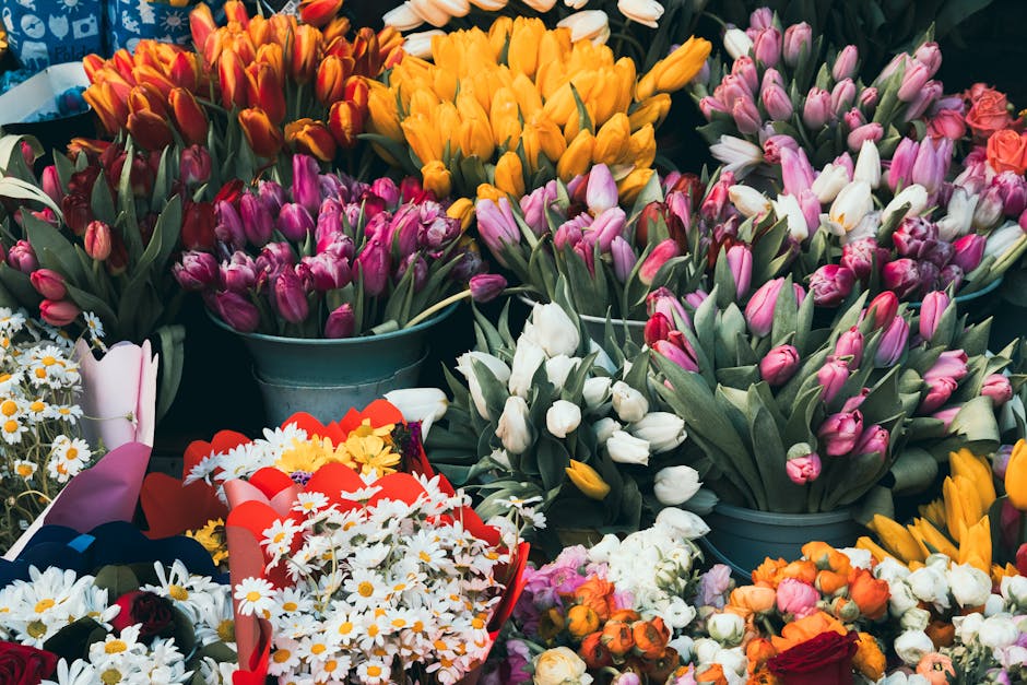 A vibrant display of fresh flower bouquets and arrangements featuring an array of tulips in shades of pink, white, orange, and yellow, arranged in metal buckets and wrapped in decorative paper with ri