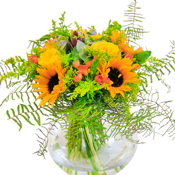 Warmth and vitality flow from this bouquet, reminiscent of golden hours in Hampton's riverside parks. Luminous sunflowers take centre stage, each one a mosaic of sunny yellow petals that seem to radiate happiness, their velvet brown centres anchoring the arrangement with confident energy. The fresh, buttery yellow of garden roses curl in perfect spirals alongside, adding textural richness and the promise of longevity-echoing the elaborate blooms cultivated in Hampton estate gardens. Hypericum berries, bright and glossy, cluster among the foliage with lively bursts of crimson-red, their sheen standing out against the smooth, matte surfaces of petals and leaves. Lush greenery cascades around the design: delicate ferns soften the edges, their feathery shapes swaying gently as if touched by a Thames breeze; sprigs of light green filler and goldenrod pepper the bouquet with a confetti of tiny yellow blossoms; eucalyptus leaves, tinged bluish-green, are quietly interspersed, enhancing the freshness. The clear, spherical vase sits serenely atop a pale surface, its transparency revealing a jungle of green stems awash in gleaming water. The scent is subtle but lively, a blend of cut grass and rose, invoking memories of open lawns and riverside walks around Hampton. Perfect for birthdays, housewarmings, or simply to uplift a friend's mood, this bouquet is a true reflection of the area's natural richness and community appreciation for floral artistry.